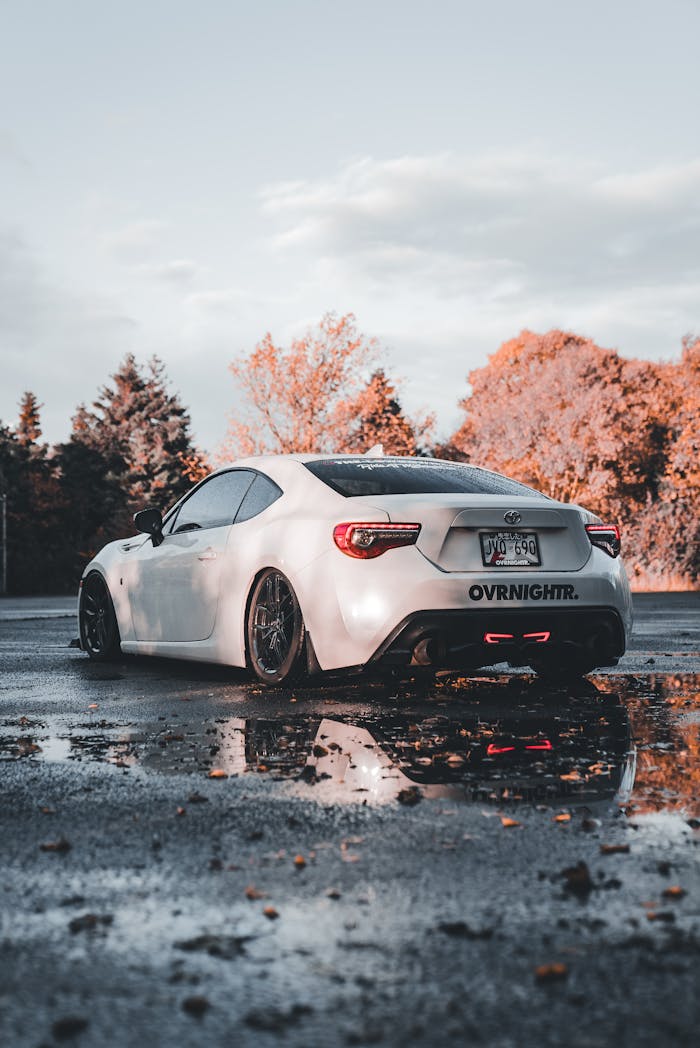 A sleek white sports car parked on a wet road with autumn trees reflecting.
